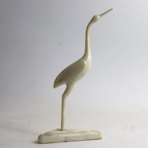 May include: A cream-colored, stylized bird figurine with a long neck and beak, standing on a rectangular base. The sculpture has a smooth, glossy finish and is set against a plain background. The bird's posture suggests a graceful, elegant design.