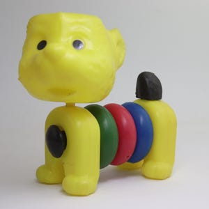 May include: A yellow plastic toy dog with a segmented body. The dog's head and legs are yellow, with black eyes and a black tail. The body segments are green, red, and blue.
