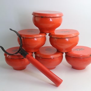 Puede incluir: Colección de seis cuencos rojos con tapa apilados en forma de pirámide. Una herramienta roja y negra con brazos curvos descansa frente a los cuencos. Los cuencos tienen un acabado brillante.