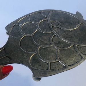 May include: A silver-colored metal trivet shaped like a fish with a textured, scaly design. The trivet has a handle and three feet.
