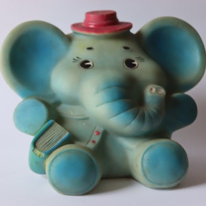 May include: A blue rubber elephant toy wearing a red hat and holding a book. The elephant has a long trunk and big ears.