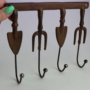 May include: Rustic metal wall hook with four hooks shaped like garden tools, including a shovel and a pitchfork. The hooks are painted in a rusty brown color and have silver ball tips.
