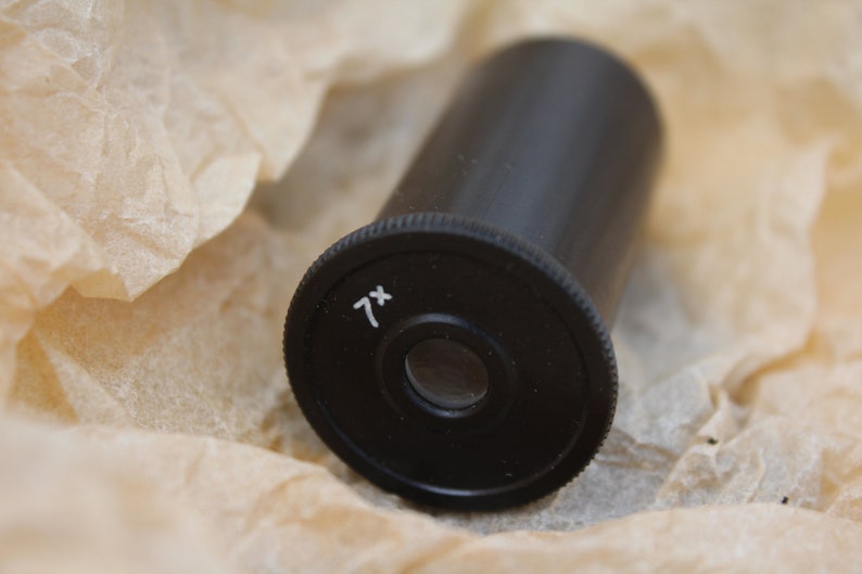 May include: A black metal magnifying lens with 7x magnification. The lens is on a brown paper background.