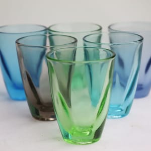 Op de afbeelding: Zes glazen shotglazen in verschillende tinten blauw en groen. De glazen zijn allemaal helder en hebben een licht gerimpelde textuur.