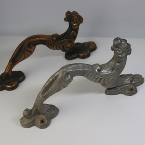 May include: Two antique door handles. One is bronze-colored with dark green accents, and the other is silver-toned. Both handles feature a decorative design with a lion's head and ornate curves. They are likely made of metal.
