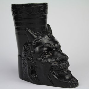 May include: Black glass figurine of a devil's head with a wide grin. The figurine is shaped like a vase or a drinking vessel.