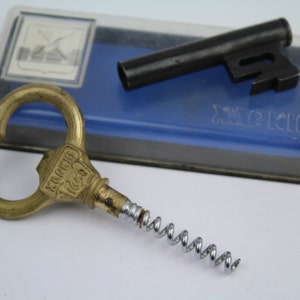 May include: A gold bottle opener with a corkscrew and a label that reads "B.P.M.  KOPECK",  next to a black metal key in a blue and clear plastic case.