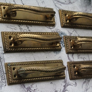 May include: Six antique brass cabinet door pulls with a rope design. Each pull has a rectangular base with a curved handle.