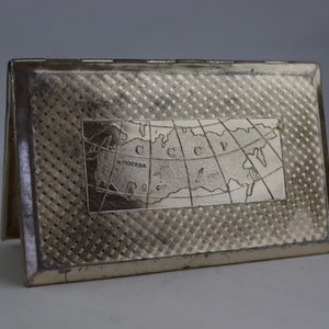 May include: A silver-coloured metal card case with a textured, cross-hatched pattern. The front has a recessed rectangular panel with a map of the USSR and the word "Moscow". The case is open, revealing the interior.