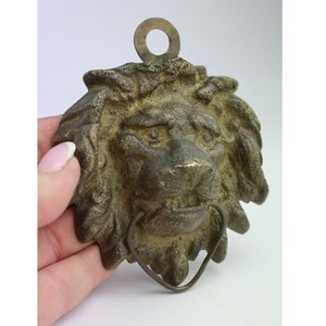 以下が含まれることがあります： 上部にリング、下部にフックが付いたブロンズ製のライオンの頭部の壁飾り。ライオンの顔は、たてがみ、目、そしてうなり声をあげている口で詳細に描かれている。