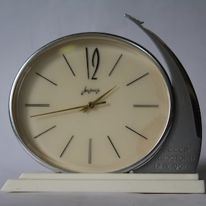 May include: A silver and white desk clock with a round face and black hands. The clock face has the word "Majak" printed on it. The clock is inscribed with "CCCP BOSTOK 12.IV.1961".