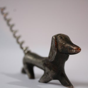 Può includere: Un apribottiglie in metallo argentato a forma di bassotto con una coda a cavatappi.