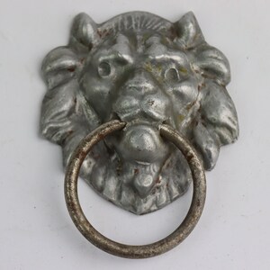 Puede incluir: Un llamador de puerta de cabeza de león plateado con un anillo redondo para tirar. La cara del león está detallada con una melena y una expresión feroz.