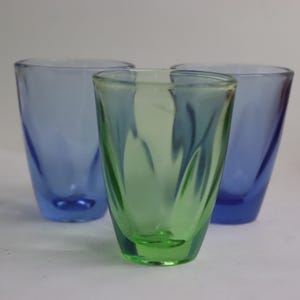 Puede incluir: Tres vasos de chupito de vidrio, uno verde y dos azules. El vaso verde tiene un diseño ondulado.