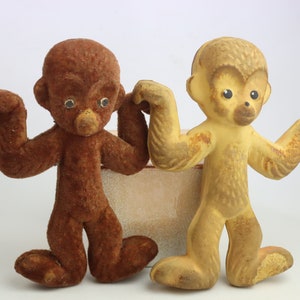 Puede incluir: Dos juguetes de monos vintage. Uno es marrón, el otro es de color canela. Ambos tienen los brazos extendidos y están en posición de pie. Los monos tienen ojos pequeños y redondos y una superficie texturizada, similar al peluche. Están colocados frente a una caja de color neutro.