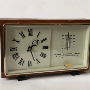 May include: A brown wooden framed clock with a white face and Roman numeral hour markers. The clock also has a thermometer with a white face and black markings. The clock is made by the Max brand.