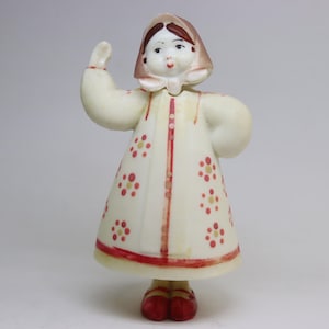 May include: A ceramic figurine of a woman in a white dress with red and yellow floral accents. The dress has a red trim and red buttons. The woman is wearing a headscarf and red shoes. She is waving with her right hand.