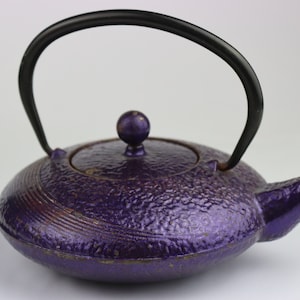 May include: A purple cast iron teapot with a black handle and a round lid. The teapot has a textured surface and a slightly curved spout.
