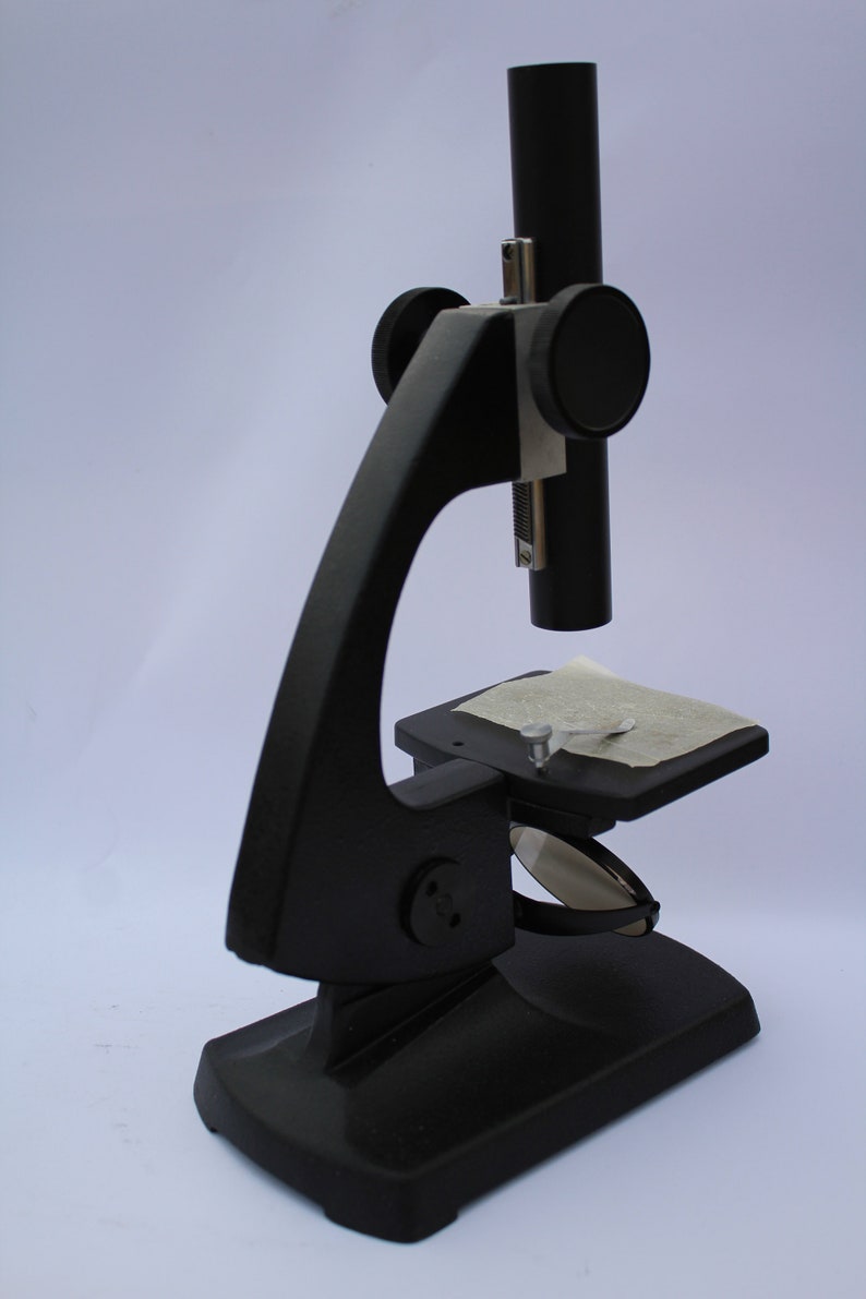 May include: A black microscope with a stage and a single lens. The microscope is on a white surface.
