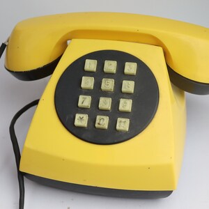 May include: A bright yellow rotary phone with a black keypad and a black cord. The phone has a simple design with a rounded shape.