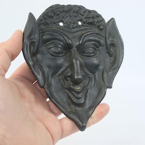 Könnte beinhalten: Eine dunkle Maske im antiken Stil mit einem teuflischen Gesicht. Die Maske hat große, spitze Ohren, eine markante Nase und eine lockige Frisur. Die Maske wird vor einem weißen Hintergrund gehalten.