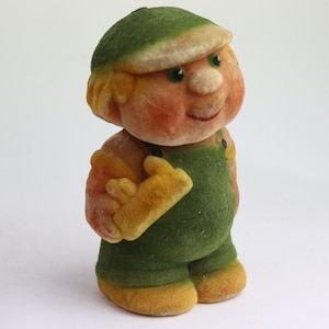 Puede incluir: Una figura de juguete de peluche de un niño que lleva un sombrero verde y un mono. El juguete tiene una cara sonriente con ojos verdes y una herramienta amarilla en la mano.