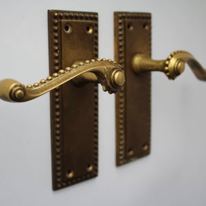 May include: A pair of antique brass door handles with a decorative beaded trim. The handles have a curved design and are mounted on rectangular plates.