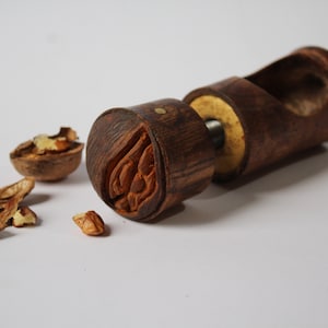 May include: A wooden nutcracker with a carved design on the top. The nutcracker is made of dark brown wood and has a metal screw in the middle. There are some walnut shells on the table next to the nutcracker.