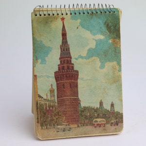 Op de afbeelding: Een vintage notitieboekje met spiraalbinding en een vervaagde afbeelding van een rode bakstenen toren met een ster erop. De toren wordt omringd door bomen en een blauwe lucht met witte wolken. Het notitieboekje staat rechtop.
