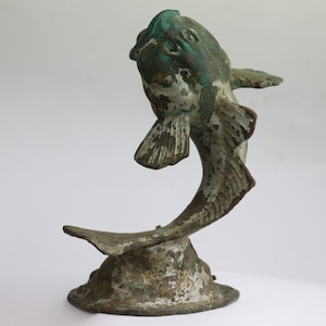 Può includere: Una scultura in metallo verde e bianco di un pesce che salta fuori dall'acqua. Il pesce è in posizione curva con la coda sollevata in alto. La scultura è su uno sfondo bianco.