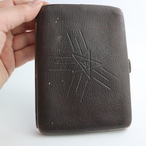 May include: A dark brown leather case with a textured surface. The case features an embossed starburst design on the front. The case is rectangular and has a clasp closure. The item is held by a hand.