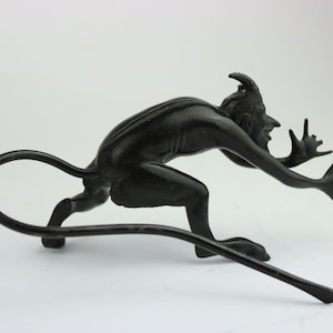 Puede incluir: Figurilla de metal negro de un diablo agachado con una cola larga y rizada y cuernos. El diablo tiene una expresión amenazante y los brazos extendidos.