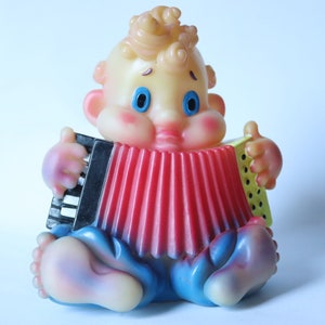 Può includere: Una figurina di giocattolo di plastica gialla di un bambino che suona una fisarmonica rossa. Il bambino ha gli occhi blu e i capelli ricci gialli.