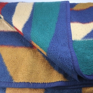 May include: A folded blanket featuring a geometric design in blue, red, tan, green, and cream. The blanket has a blue border and a textured surface. Ideal for home use or outdoor activities.