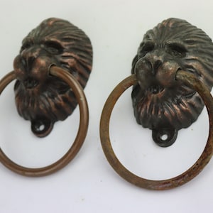 Puede incluir: Dos tiradores de puerta de cabeza de león antiguos con un acabado de bronce rústico. Los anillos son de metal y tienen una forma circular.