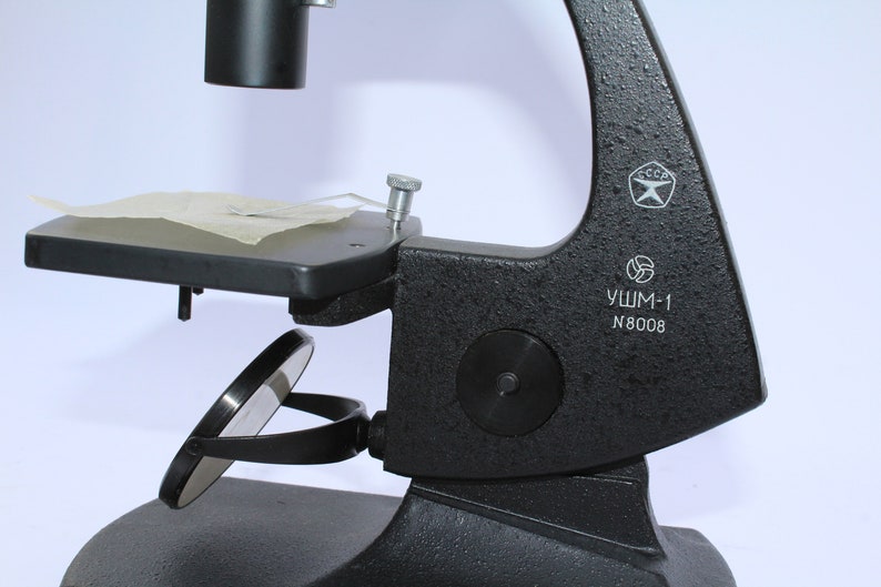 May include: A black microscope with a mirror and a stage. The microscope has a label that reads "УШМ-1 N8008" and a USSR symbol.