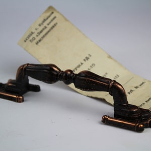 Könnte beinhalten: Ein antiker, verzierter, kupferfarbener Türgriff mit einem dekorativen Design. Der Griff liegt auf einem Stück Papier mit Text in einer Fremdsprache.