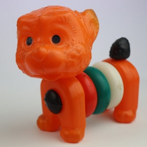 May include: Orange plastic toy dog with black eyes and a ring of colorful rings around its body.