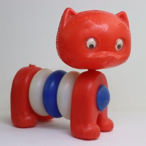 Könnte beinhalten: Eine rote und weiße Plastikspielzeugkatze mit blauen Augen und einem Stapel bunter Ringe am Körper. Die Katze ist ein Ziehspielzeug und die Ringe können gestapelt und entstapelt werden.