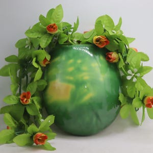 May include: A green ceramic vase with a mottled finish is decorated with a garland of green leaves and small orange and yellow flowers.