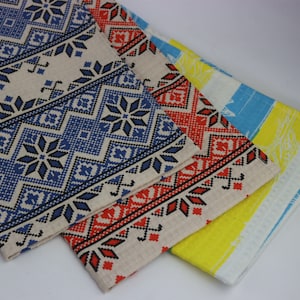 May include: Three kitchen towels with different patterns. The top towel has a blue and black geometric pattern on a beige background. The middle towel has a red and black geometric pattern on a beige background. The bottom towel has a yellow and blue striped pattern with a white floral design.