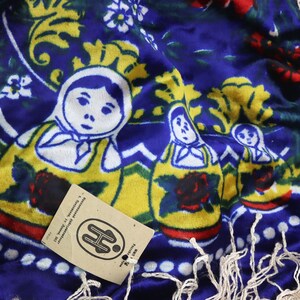 May include: A blue fabric scarf with a fringe border features a repeating pattern of yellow, red, and white Russian nesting dolls. The scarf has a small brown tag with the letter "H" in white.