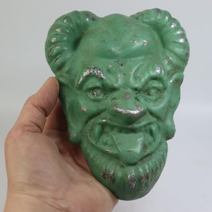 May include: A green devil face sculpture with horns and a grimacing expression. The sculpture has a distressed appearance with silver accents, suggesting it is vintage or antique. The sculpture's tongue is protruding.