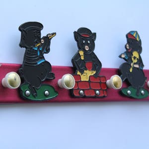 May include: A red wooden coat rack with three black pig figurines. The pig on the left is playing a trumpet, the middle pig is holding a brick, and the pig on the right is holding a paintbrush.