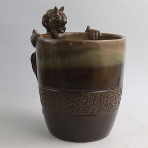 Puede incluir: Una taza de cerámica marrón con una banda texturizada y una figura de ardilla esculpida en el borde. La taza tiene un degradado de colores, que va del marrón oscuro a un marrón y verde más claro.