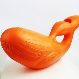 May include: Orange plastic whale toy with a spout on the top. The whale has a textured surface and a detailed design.