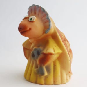 May include: A vintage, whimsical figurine of a creature with a bird-like beak and a yellow dress. The figure has a peach-colored face and body, with green eyes and a blue and peach headdress. It holds a bouquet.
