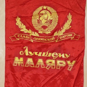 Op de afbeelding: Een rode vlag met gouden letters en een gouden embleem. Het embleem toont een krans en een ster. De tekst op de vlag luidt "Glorie aan de Leninistische Partij" en "Aan de Beste Schilder".