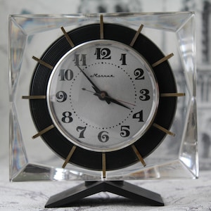 May include: A square, clear glass clock with a black face and gold accents. The clock face has black Roman numeral hour markers and black minute markers. The clock hands are black and gold. The clock is on a black stand with a triangular base. The clock face says "Маяк" in Cyrillic.
