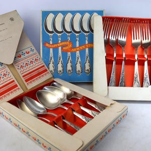 Könnte beinhalten: Ein Set aus silberfarbenen Metalllöffeln und Gabeln in der Originalverpackung. Die Löffelschachtel hat einen blauen Deckel mit einer Löffelgrafik und rotem Text, während das Innere Löffel mit verzierten Griffen zeigt. Die Gabelschachtel ist rot.